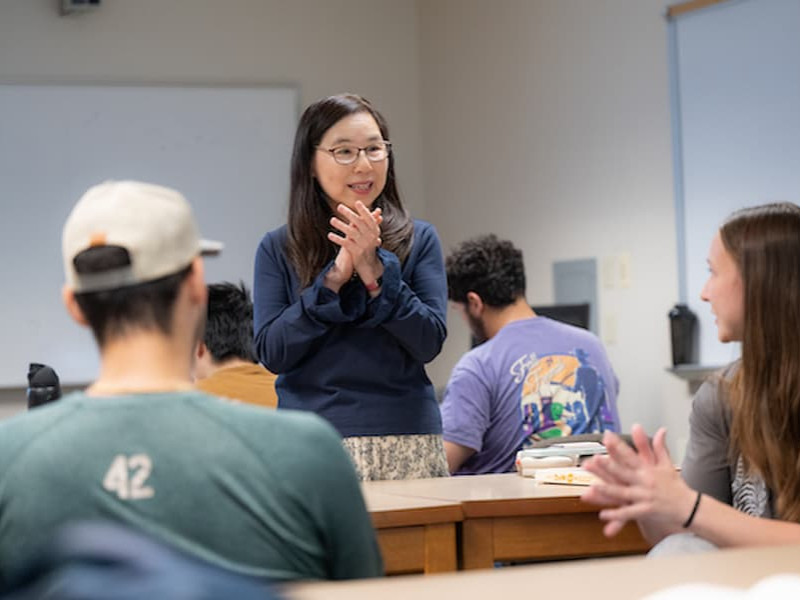 A female professor speaking and gesturing with her hands in front of students during a university classroom lecture.