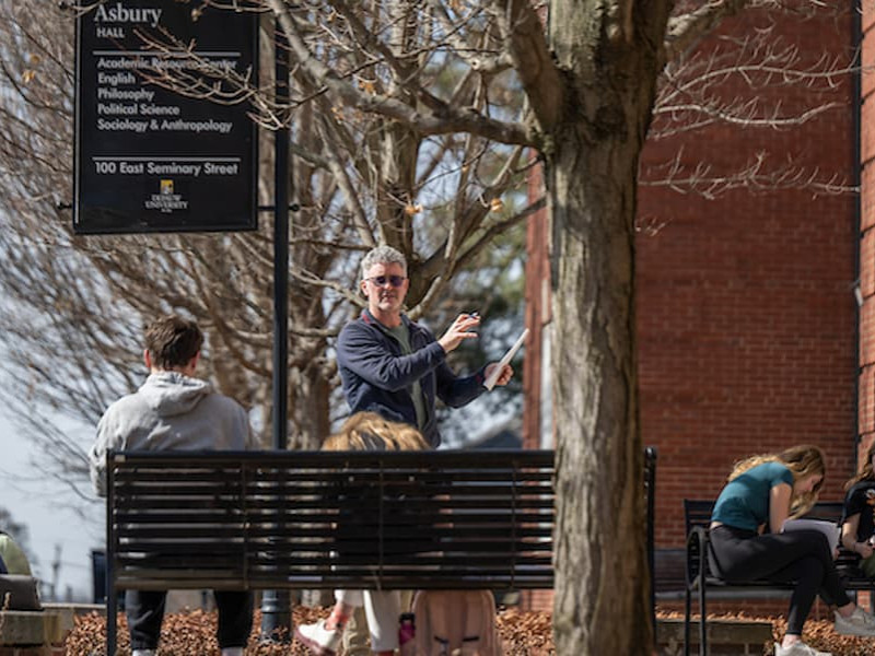 A professor standing and lecturing to students near an outdoor bench, with an Asbury Hall campus sign visible in the background.