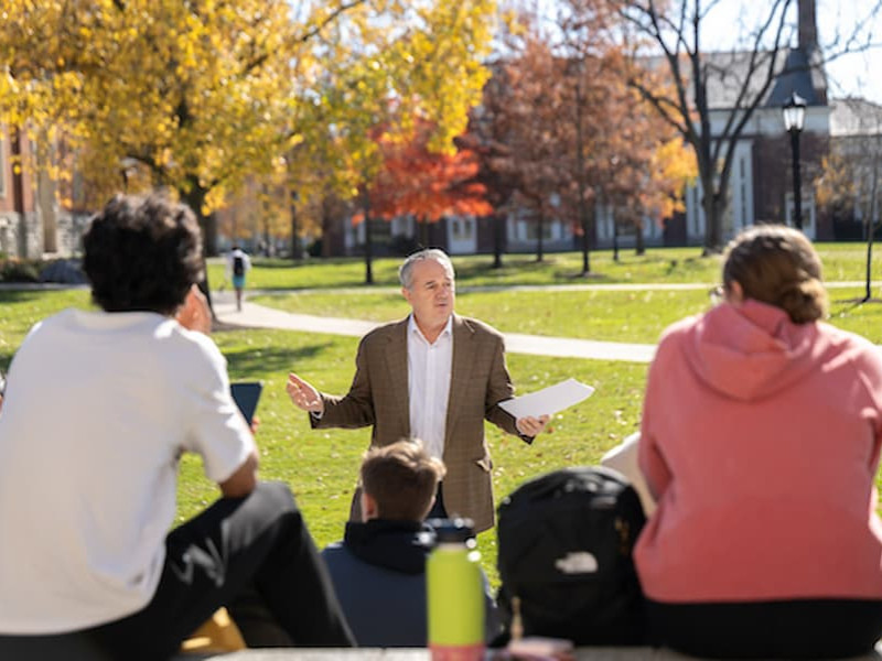 A professor holding papers and gesturing while leading a class discussion with students seated outdoors on a grassy campus lawn.