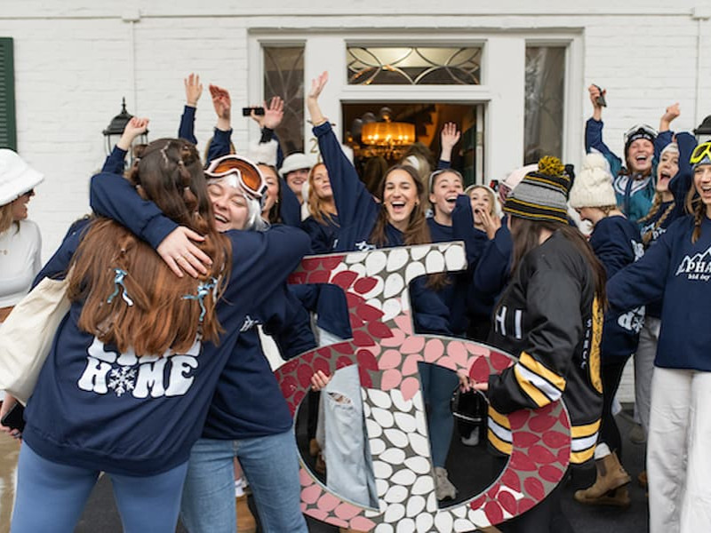 Students celebrate sorority bid day outside a brick house, hugging and cheering while holding large Greek letters.