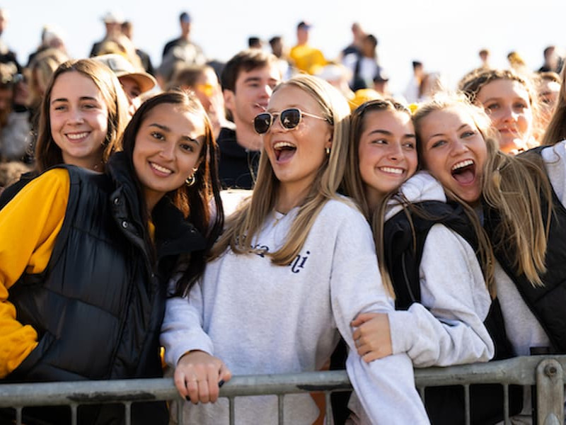 A group of smiling and cheering college students leaning against a railing in the bleachers at an outdoor sporting event.