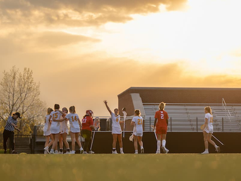 Lacrosse players in white uniforms huddle and raise their arms in celebration during a game at sunset.