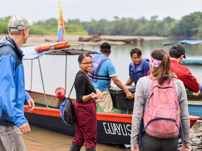 A smiling young woman looks back while preparing to board a small boat with a group of travelers and guides on a riverbank.