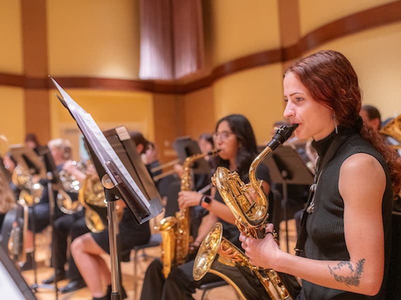 A young woman plays a saxophone on stage during an indoor concert with an orchestra ensemble.