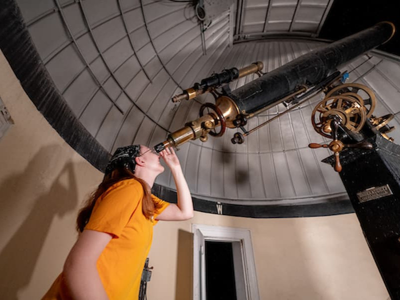 A student looks through the eyepiece of a large, vintage refracting telescope inside an observatory dome.