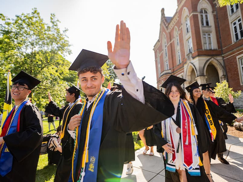 Graduating students in caps and gowns smiling and waving during a commencement procession outside historic campus buildings.