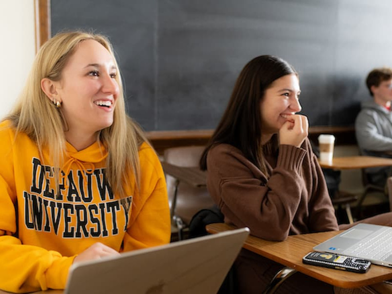 Two smiling students sitting at classroom desks with laptops, with one student wearing a yellow DePauw University hoodie.