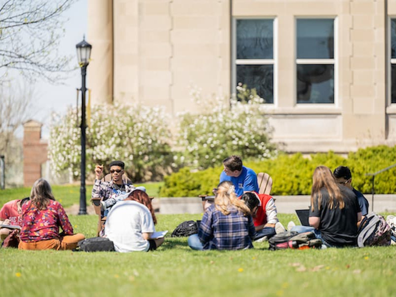 A group of students sitting in a circle on a grassy campus lawn, studying and talking together outside a stone building.