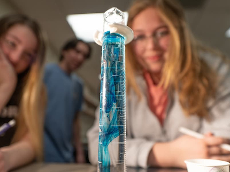 Students watch closely as blue liquid swirls inside a graduated cylinder during a science lab experiment.