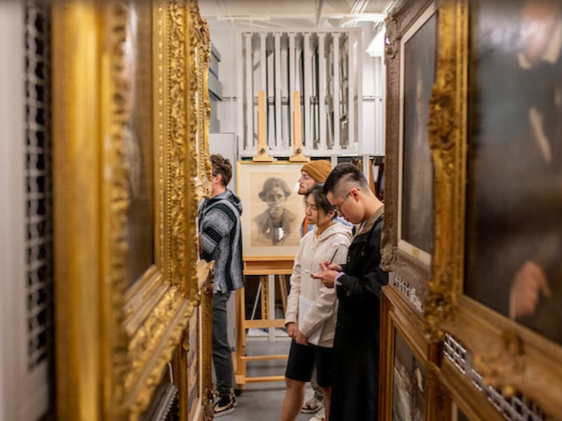 Students stand in a narrow museum archive storage room, studying a framed portrait on an easel while surrounded by racks of historical paintings.