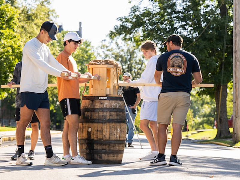 A group of male students operates a wooden capstan winch mechanism attached to a large barrel outdoors on a sunny campus street.