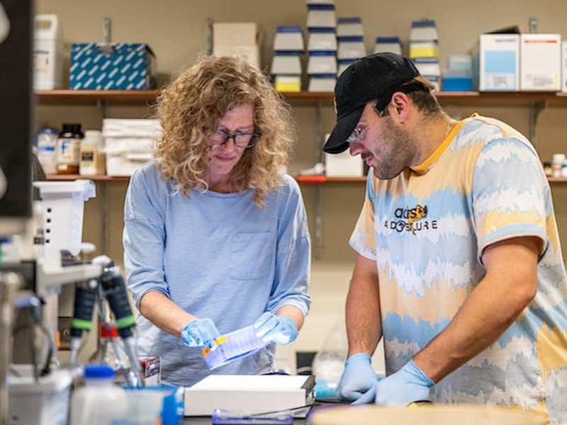 An instructor and a student wearing gloves and safety glasses work together with lab equipment in a science laboratory.