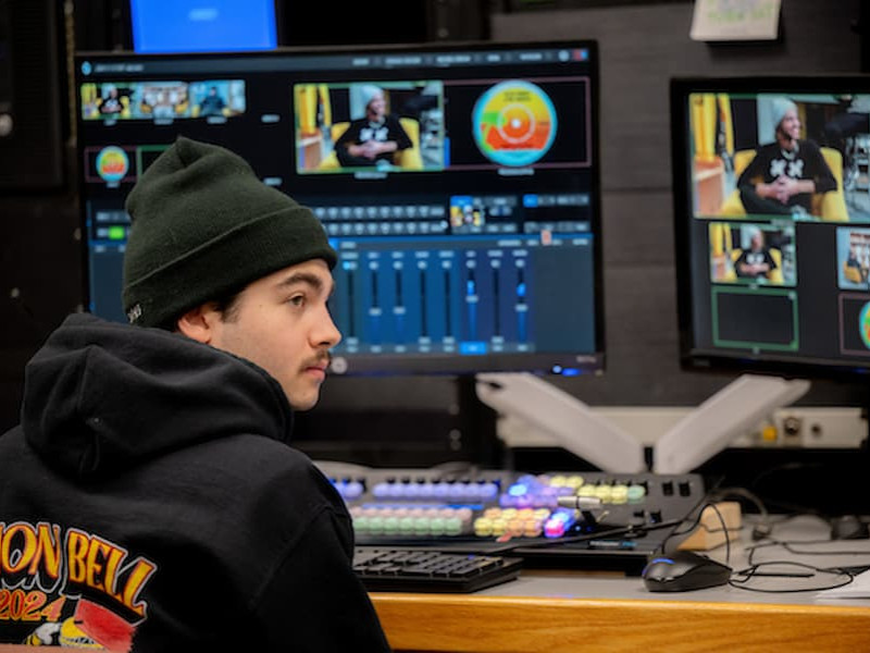 A student wearing a beanie sits at a broadcast control board, monitoring video feeds on multiple screens in a production studio.