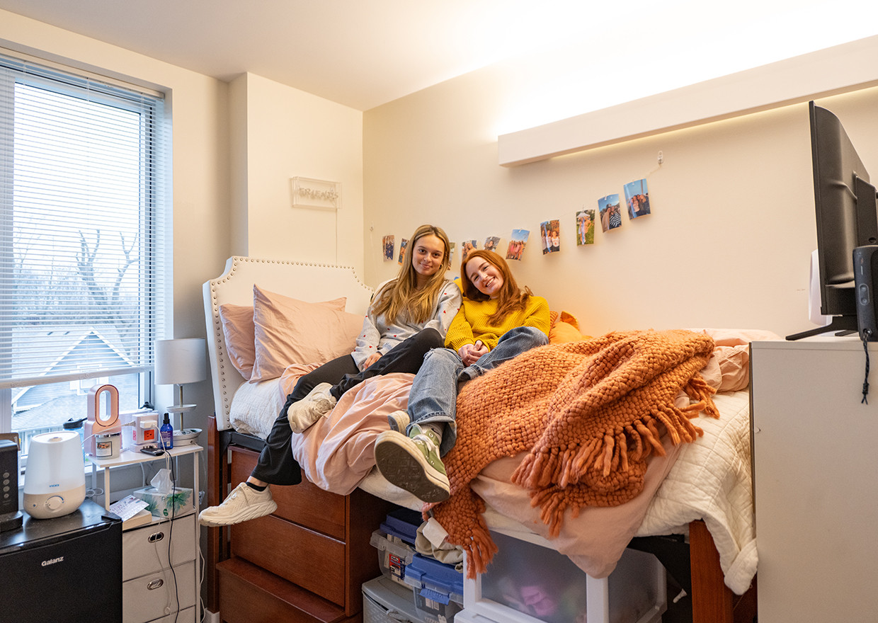 Two female students smiling as they sit together on a bed in their dorm room.