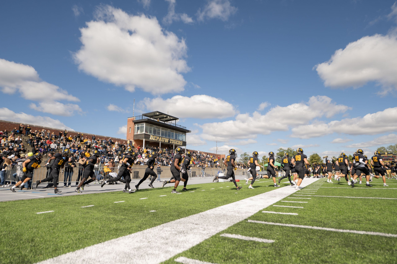 DePauw's football team runs across the field with cheering fans in the stands of Blackstock Stadium behind them.