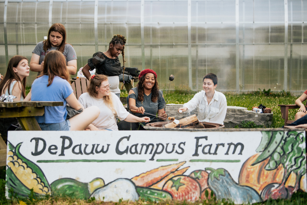 Group of students laughing and talking while roasting marshmallows on a bonfire behind the Ullem Campus Farm sign.