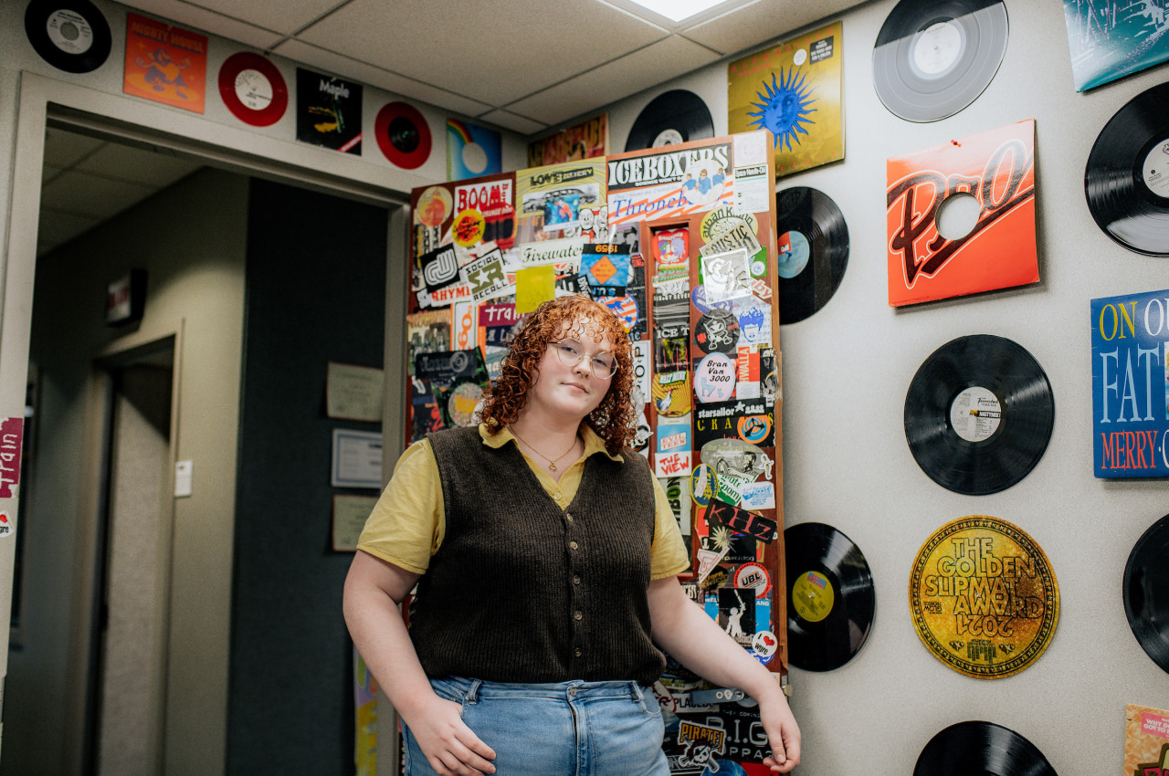 Female student posed while opening a door covered in stickers to a room with records lining the walls.