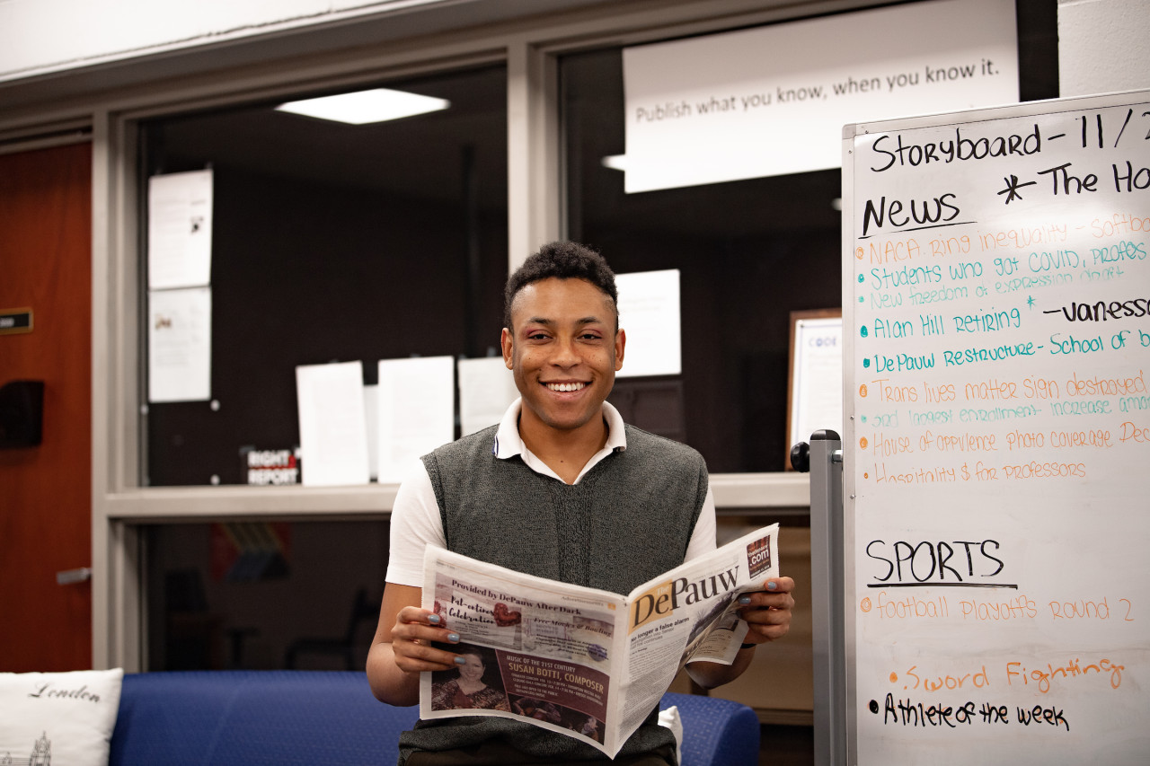 Male student smiling as he looks up from a DePauw Newspaper with a storyboard written on a whiteboard behind him.
