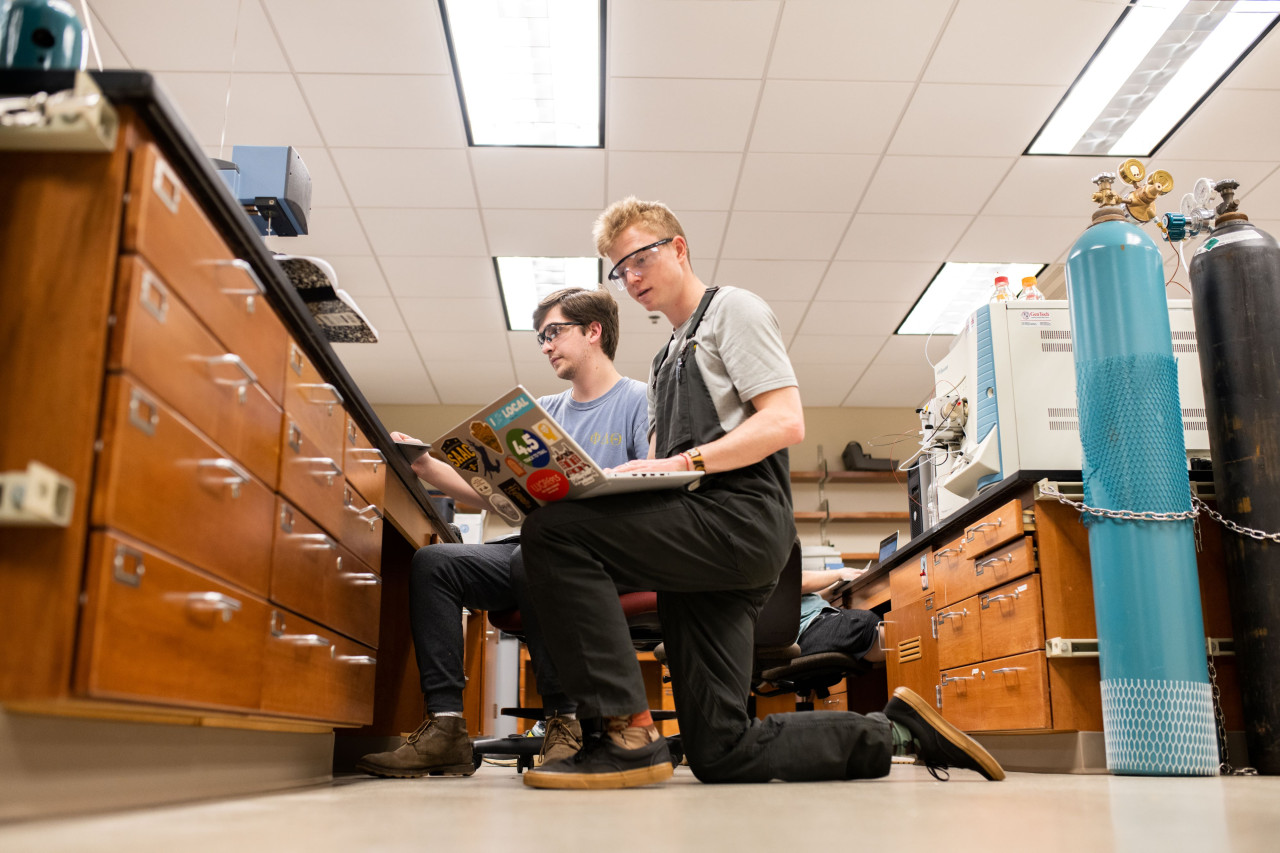 Two male students kneel between lab tables in the Organic Lab to make a note on a laptop.