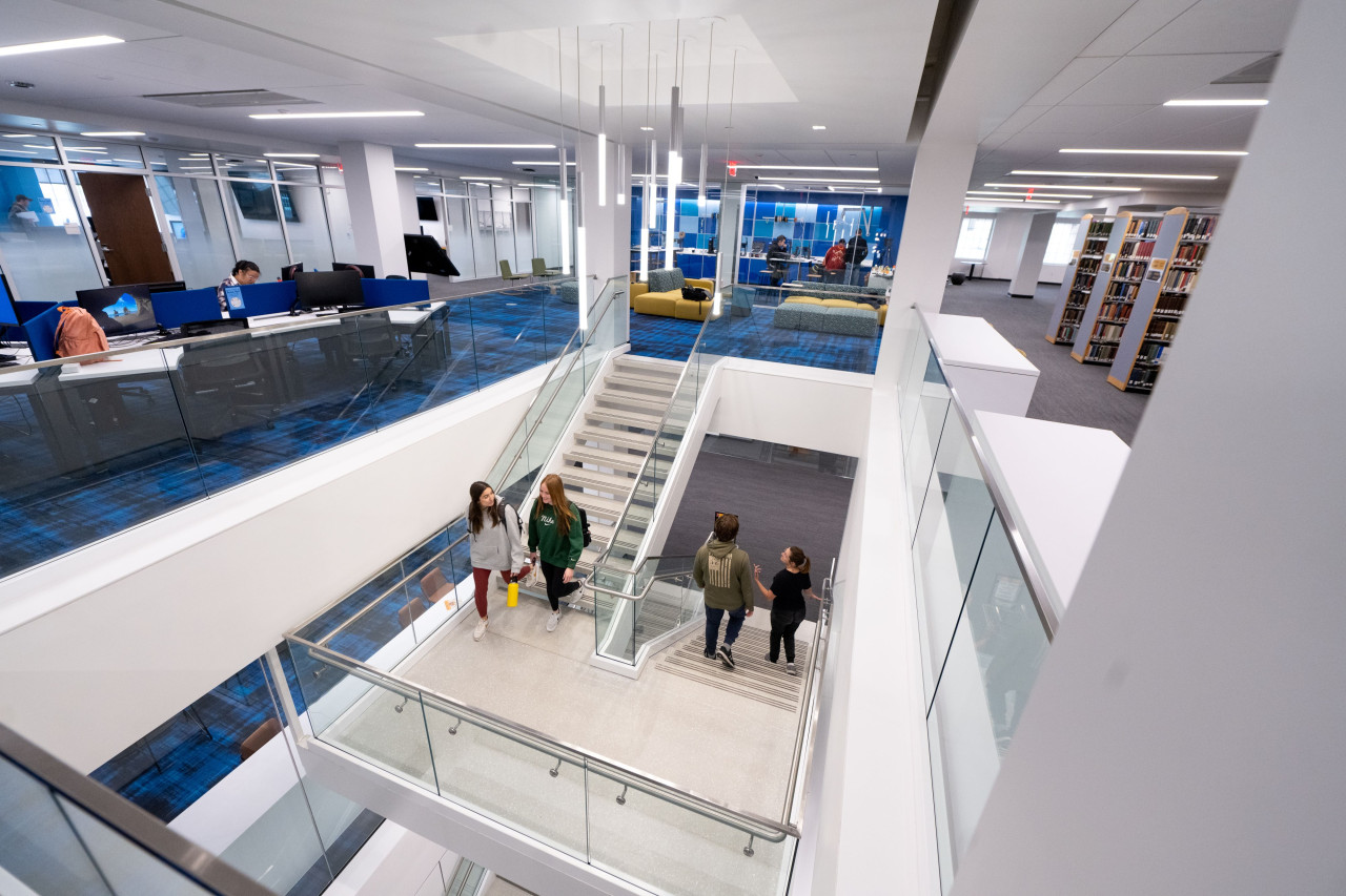 Wide lens view of students walking down staircase from a second floor with study area seating on left, technology center in back and bookcases to the right.