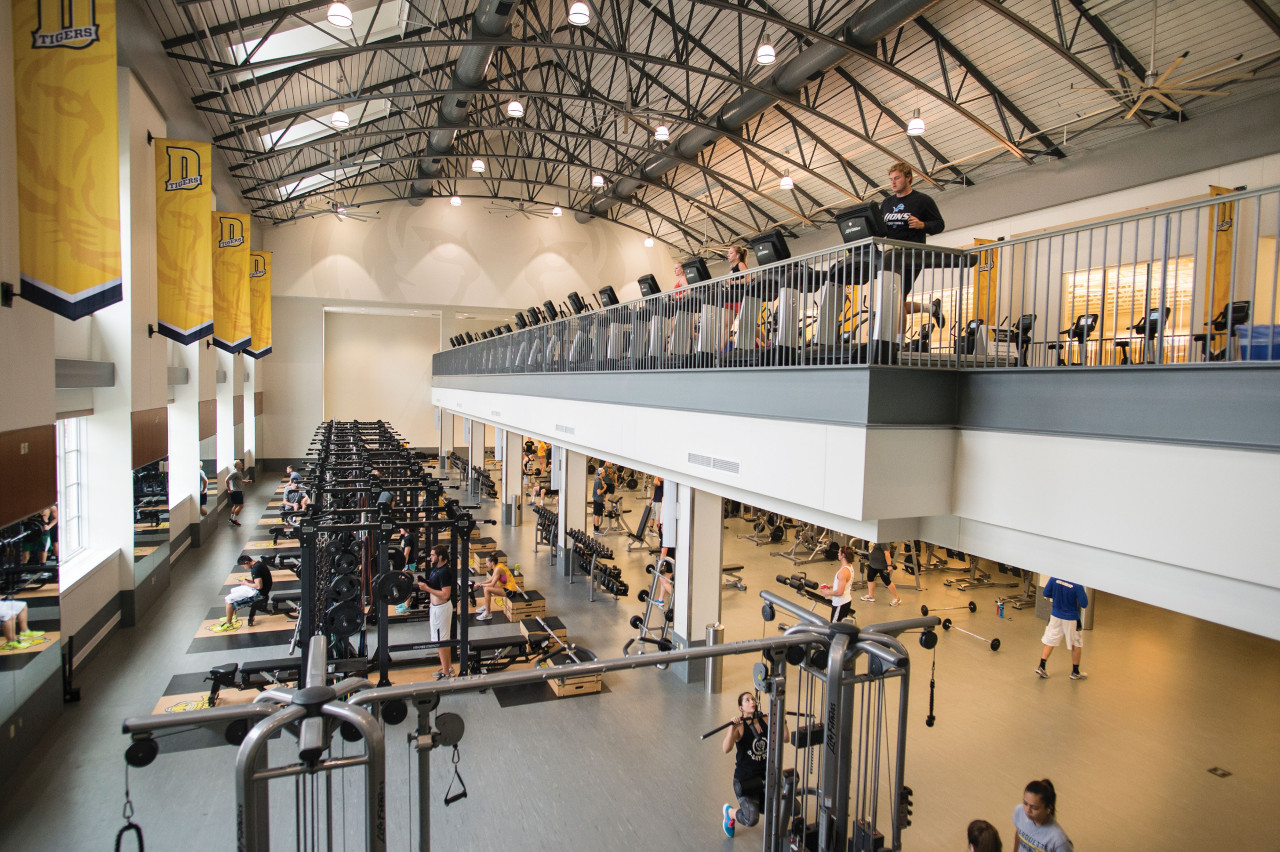 Birds eye view of students working out in Welch Fitness Center with a line of weight racks on the first floor and a line of cardio machines on the mezzanine level