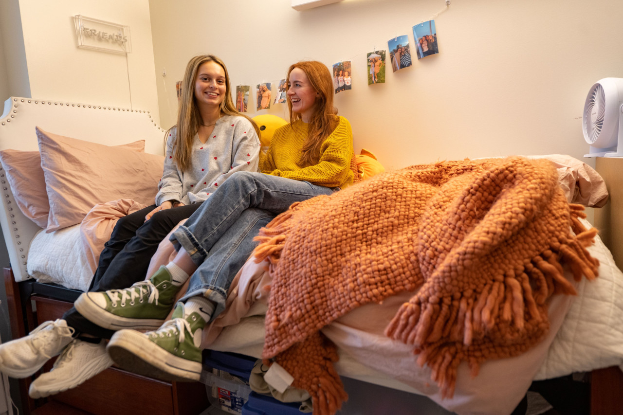 Two female students smiling as they sit together on a bed in their dorm room.